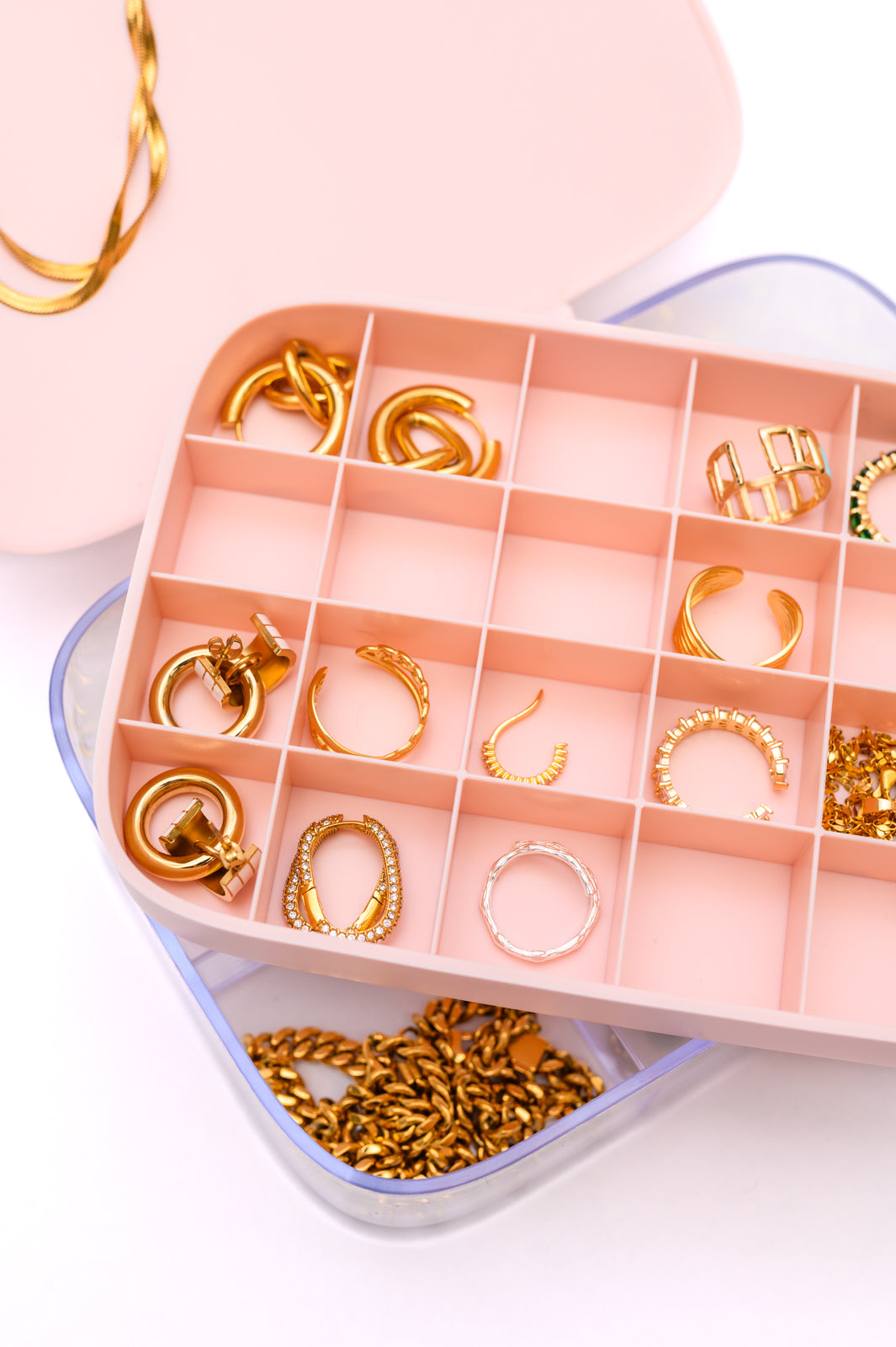 A close-up image of The Dapper Squirrel's All Sorted Out Jewelry Storage Case in Pink showcases an elegant organizational solution. The box features multiple compartments, each holding different types of gold jewelry, including earrings, rings, and chains in various designs. The removable storage tray sits neatly inside the slightly open lid.