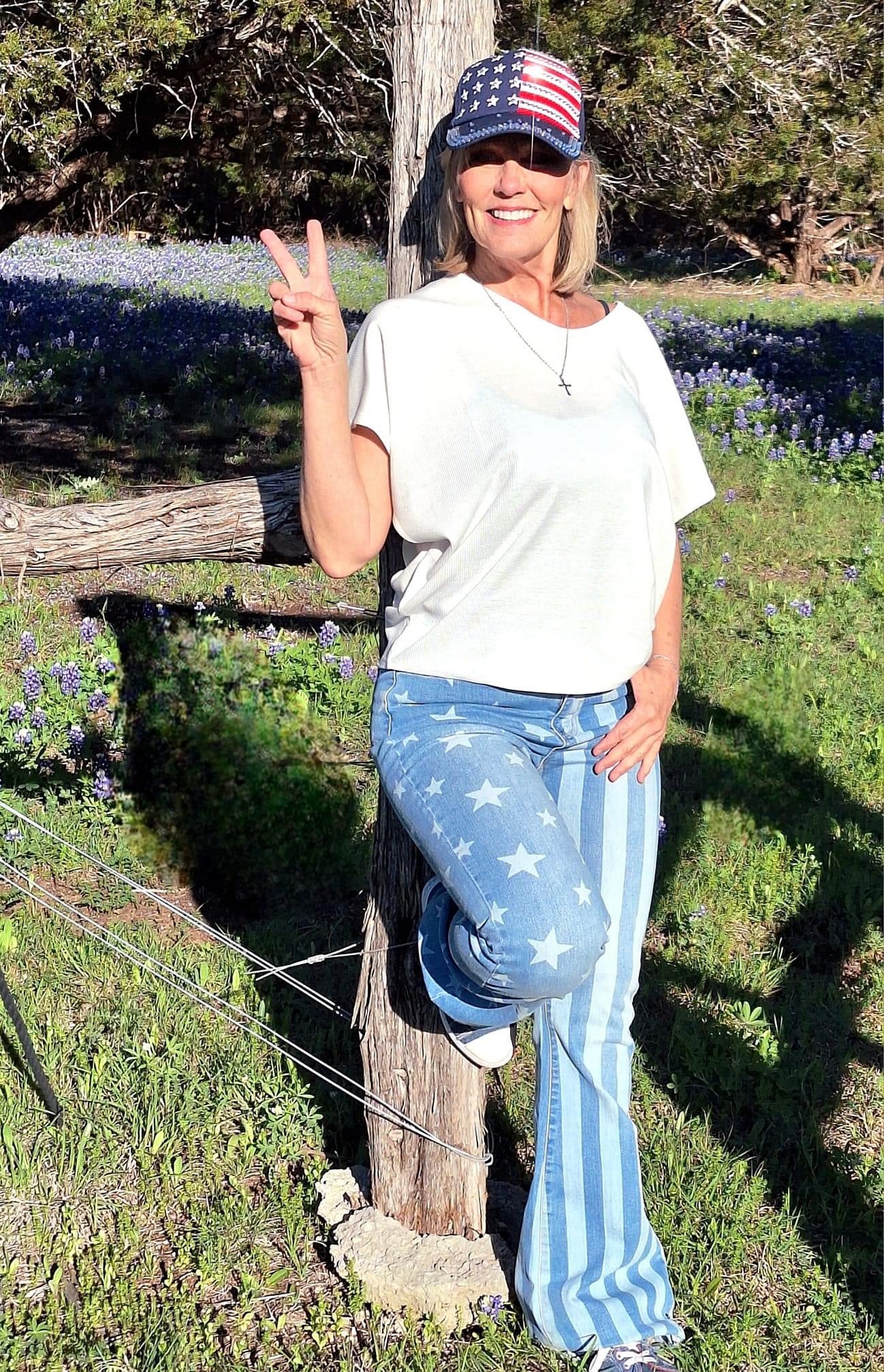 A woman wearing a white T-shirt, Freedom Rings - Judy Blue Flares by JB Boutique Simplified, and a cap with an American flag design stands outdoors by a wooden post in a field of wildflowers. She is smiling and making a peace sign with her hand.