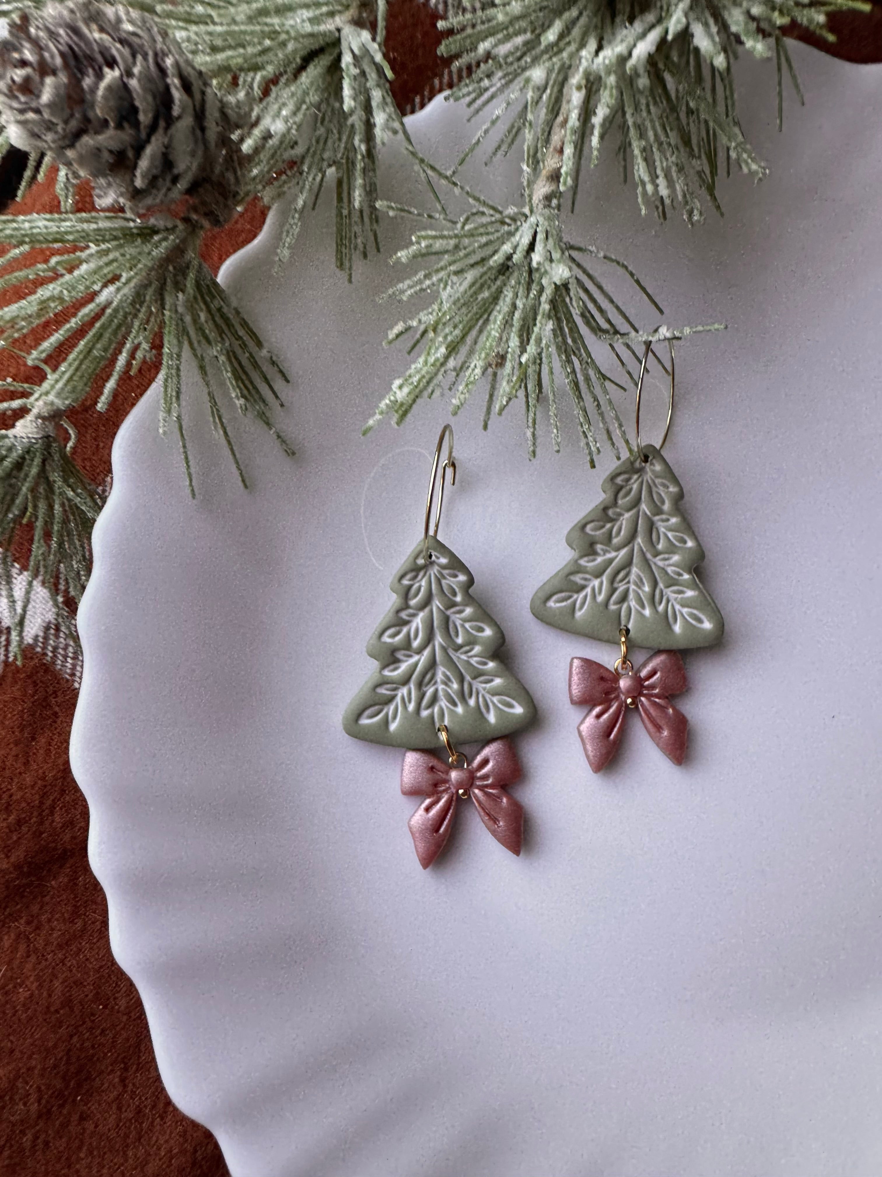 Christmas-themed earrings with green tree shapes and pink bows on a white plate with pine branches.