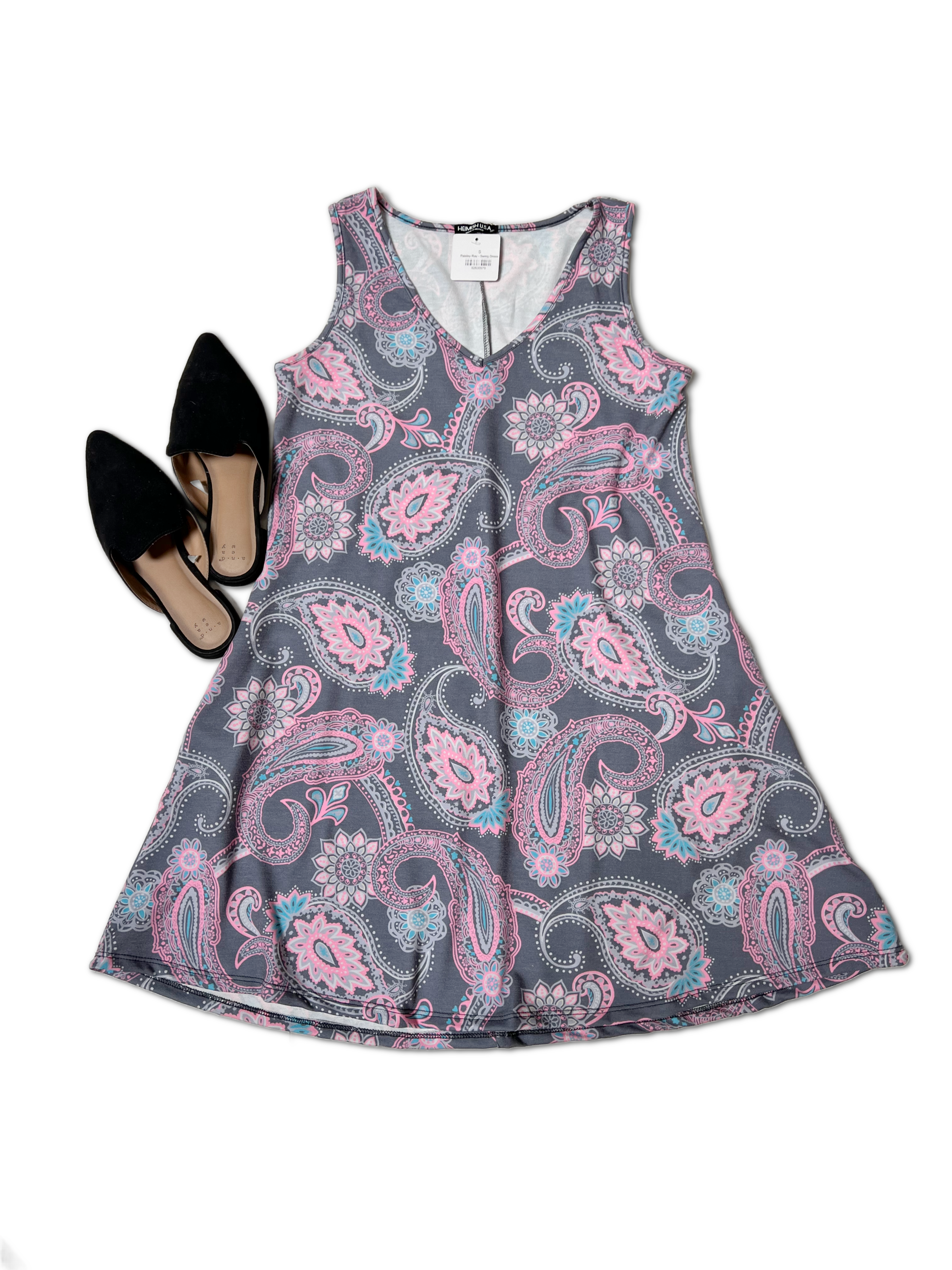 A sleeveless V-neck A-line silhouette dress with a pink and blue paisley print on a gray background is laid flat. To the left of the Paisley Ray - Swing Dress by Heimish, there is a pair of black pointed-toe flats. The image is set against a white background.