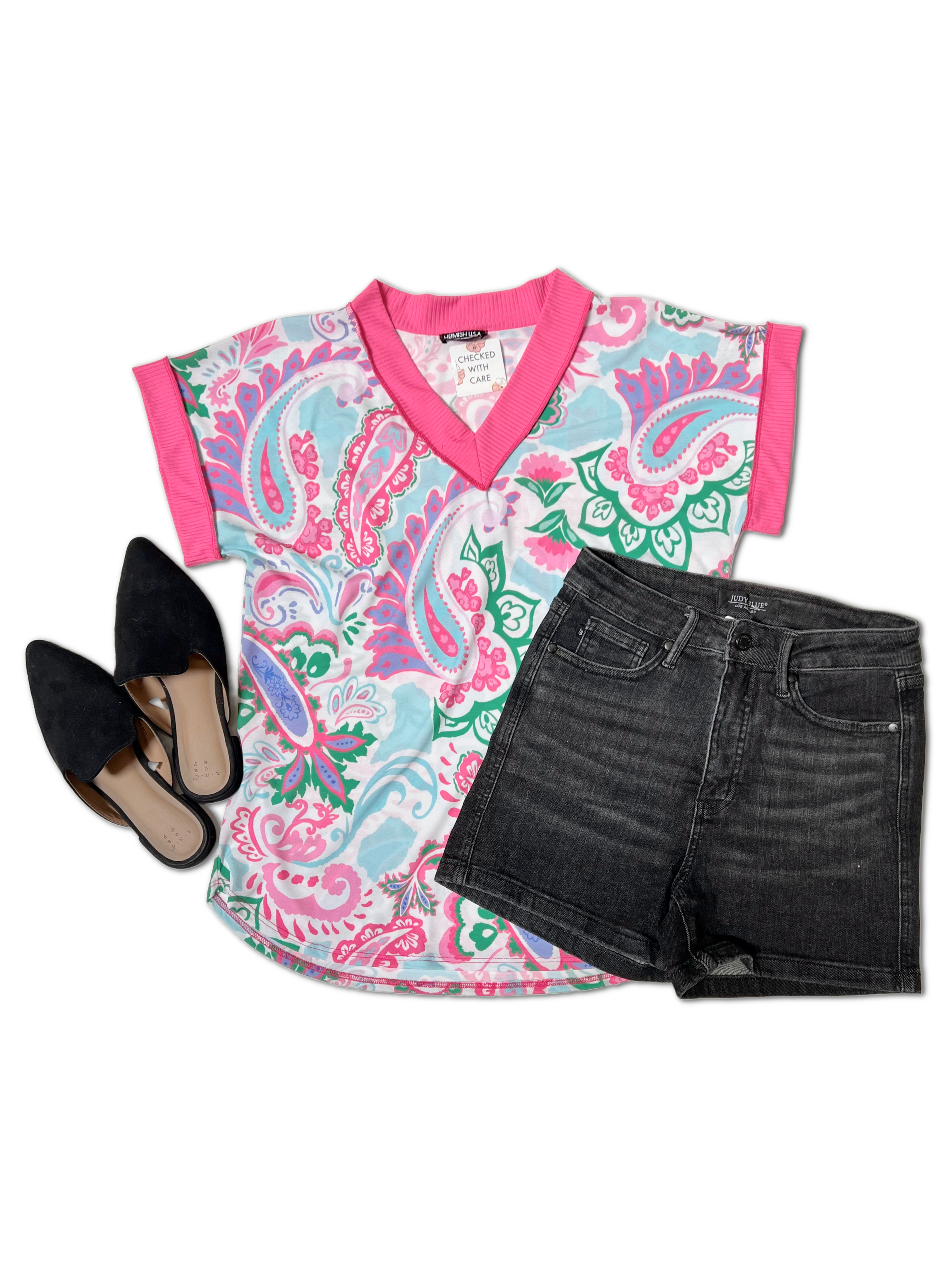 A brightly patterned Electric Paisley - Short Sleeve shirt by Heimish with pink trim is laid out on a white background. Next to it, there are black denim shorts and a pair of black, pointed-toe slide shoes. The short sleeve shirt features a mix of pink, teal, and white paisley designs.