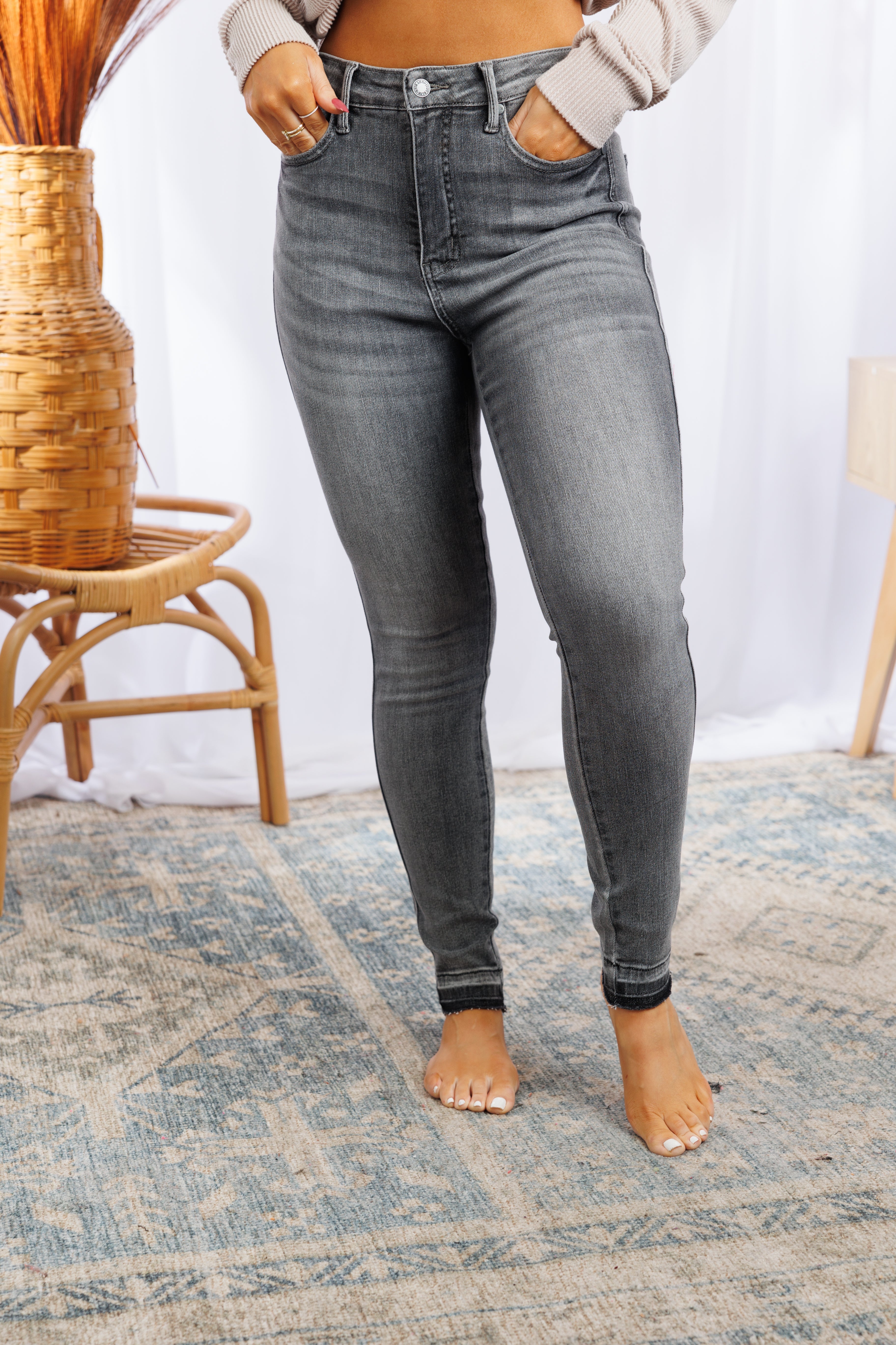 A person wearing JB Boutique Simplified's Total Control - Tummy Control Jeans with a beige top stands barefoot on a patterned rug. The surroundings include a wicker basket with dried plants and a wooden chair, all set against a backdrop of sheer white curtains. The photo focuses on the lower body.