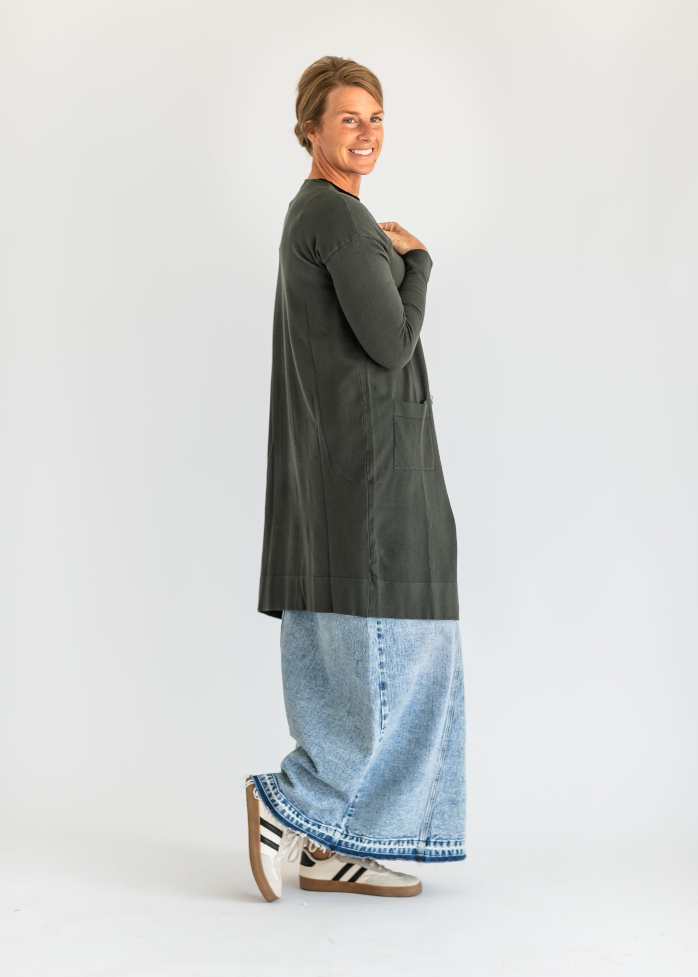 Person wearing a long dark green cardigan over a light blue denim dress on a white background