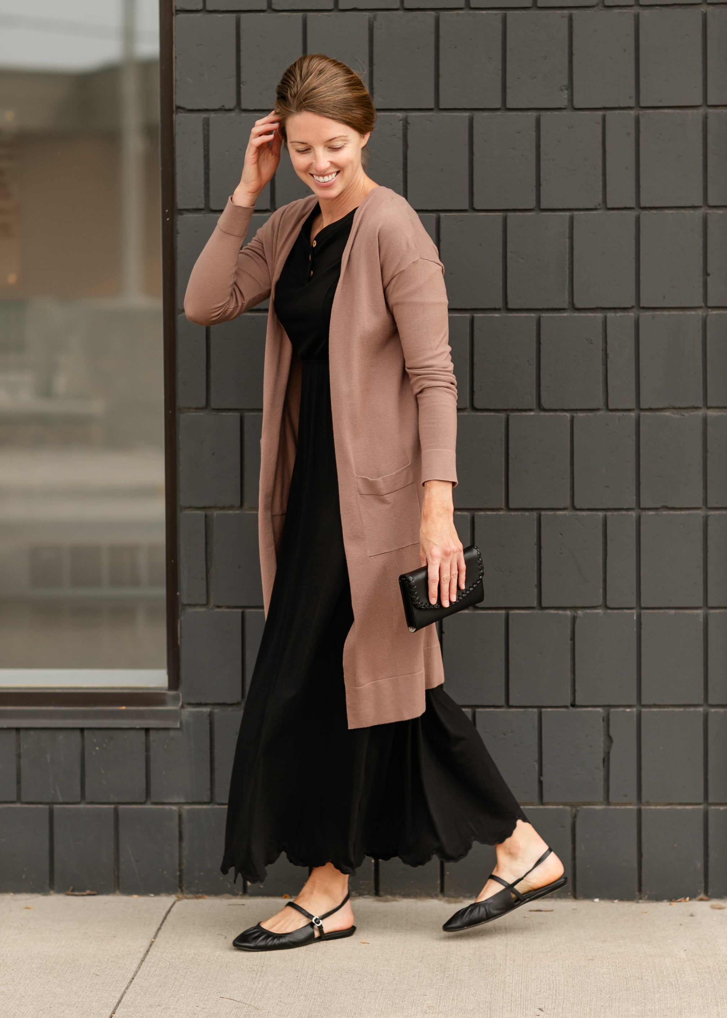 Woman wearing a long beige cardigan over a black dress against a black tiled wall.