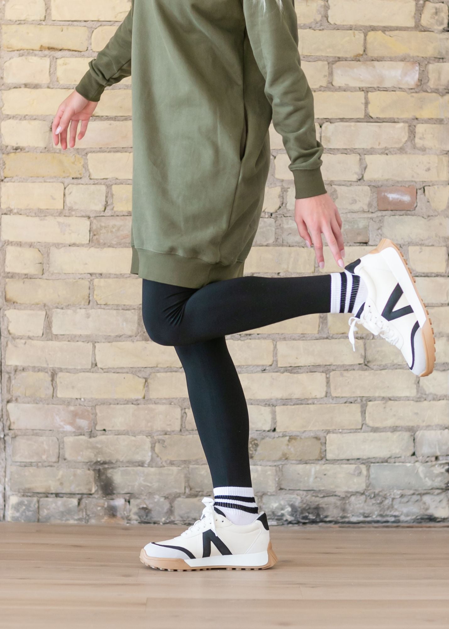 Person wearing a green dress, black leggings, and white sneakers against a brick wall.
