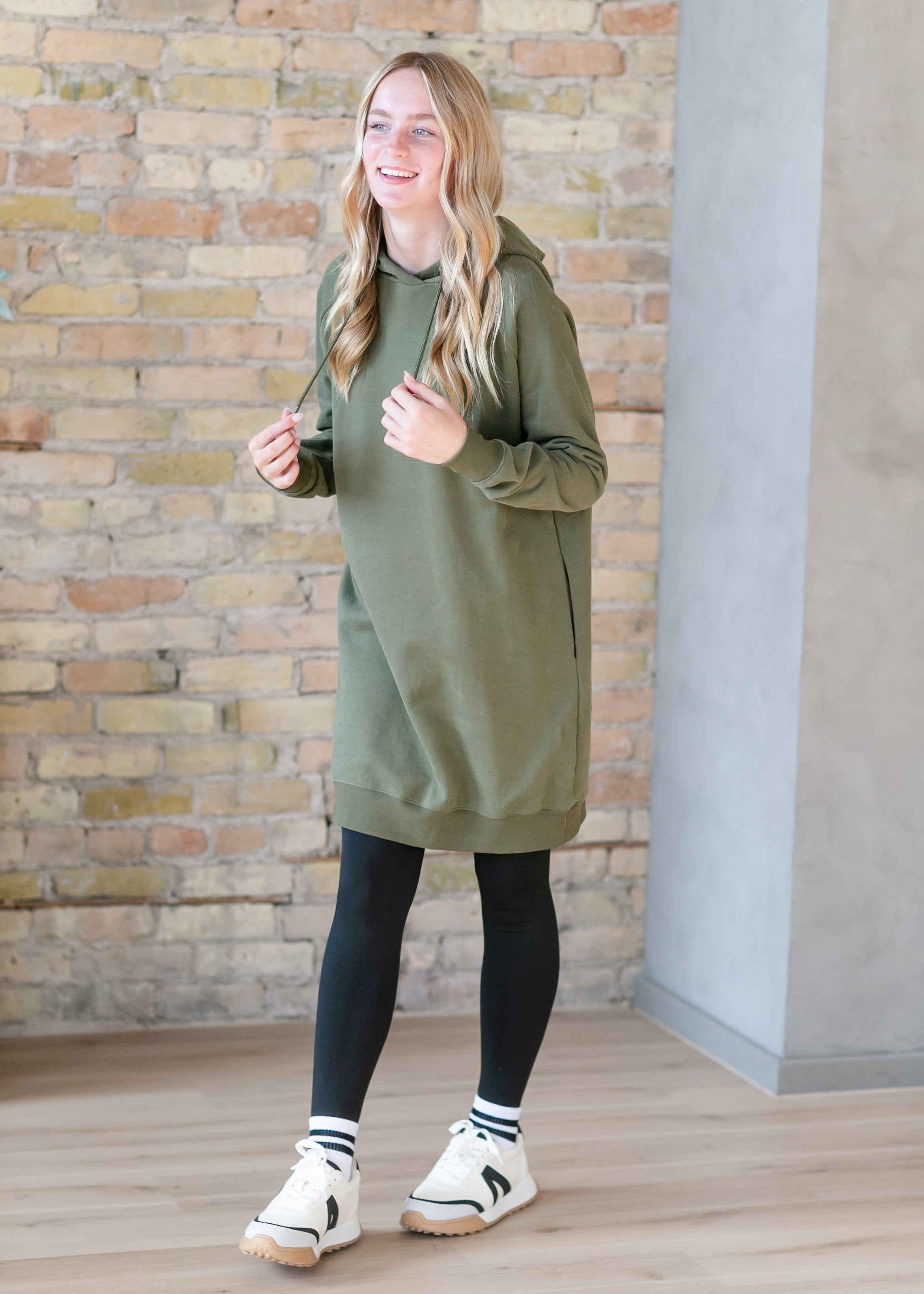 Woman wearing an olive green dress with black leggings and white sneakers against a brick wall.