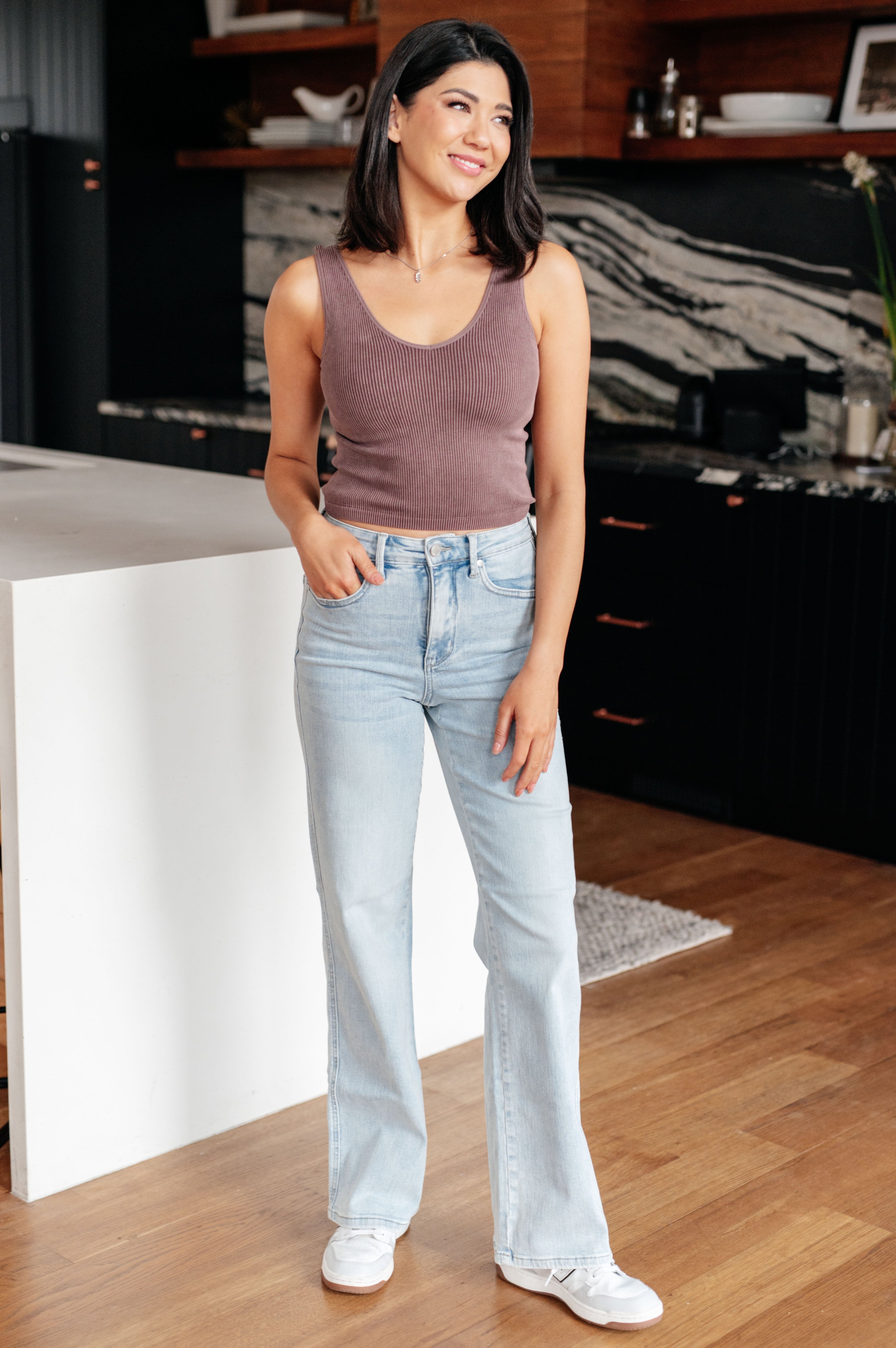 A person with long brown hair is standing in a modern kitchen, wearing a black sleeveless top and light blue Brooke High Rise Control Top Vintage Wash Straight Jeans by Judy Blue with one hand in their pocket. The kitchen features dark cabinets, a white island, and wooden floors.