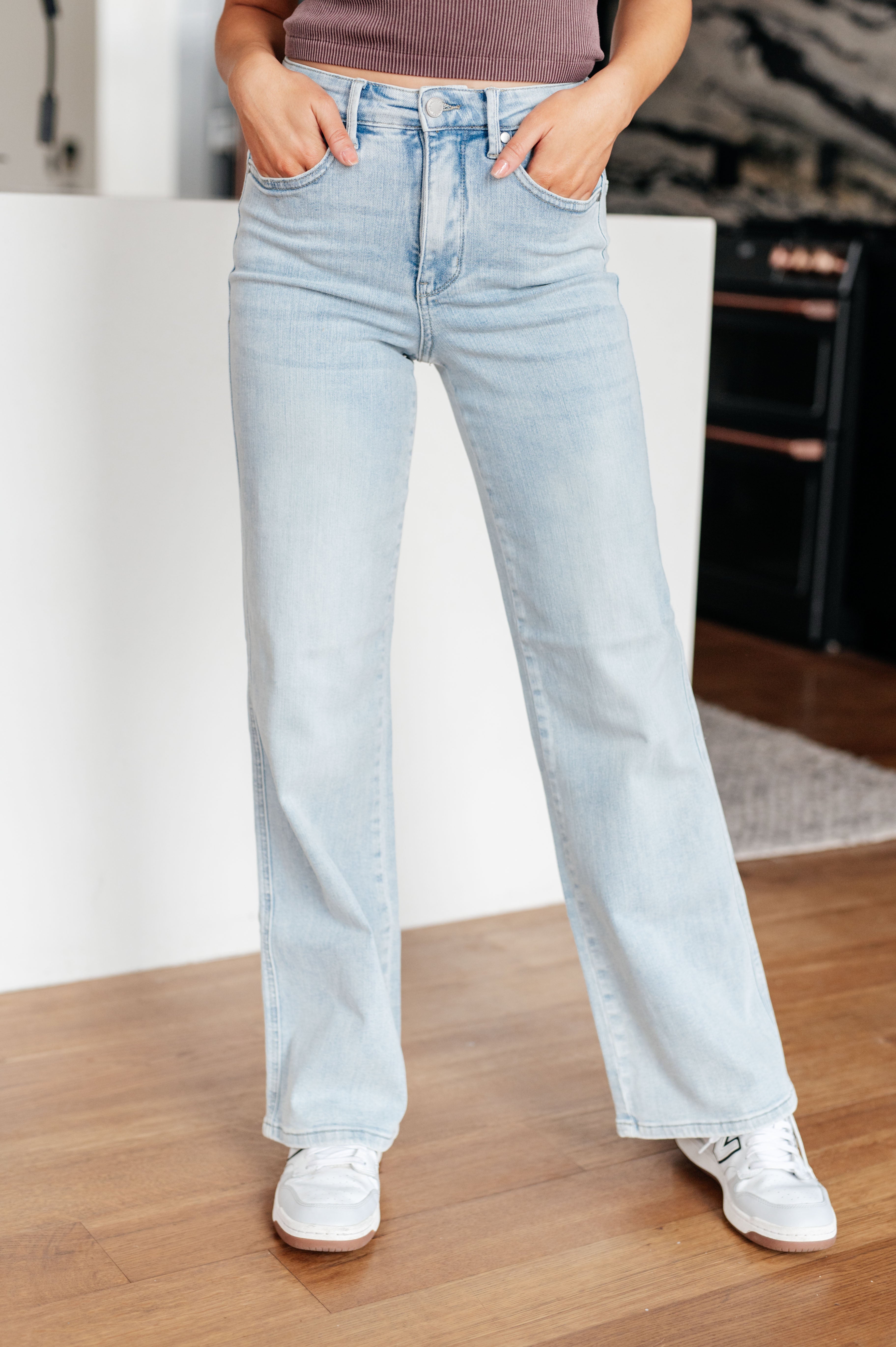 A person with long brown hair is standing in a modern kitchen, wearing a black sleeveless top and light blue Brooke High Rise Control Top Vintage Wash Straight Jeans by Judy Blue with one hand in their pocket. The kitchen features dark cabinets, a white island, and wooden floors.
