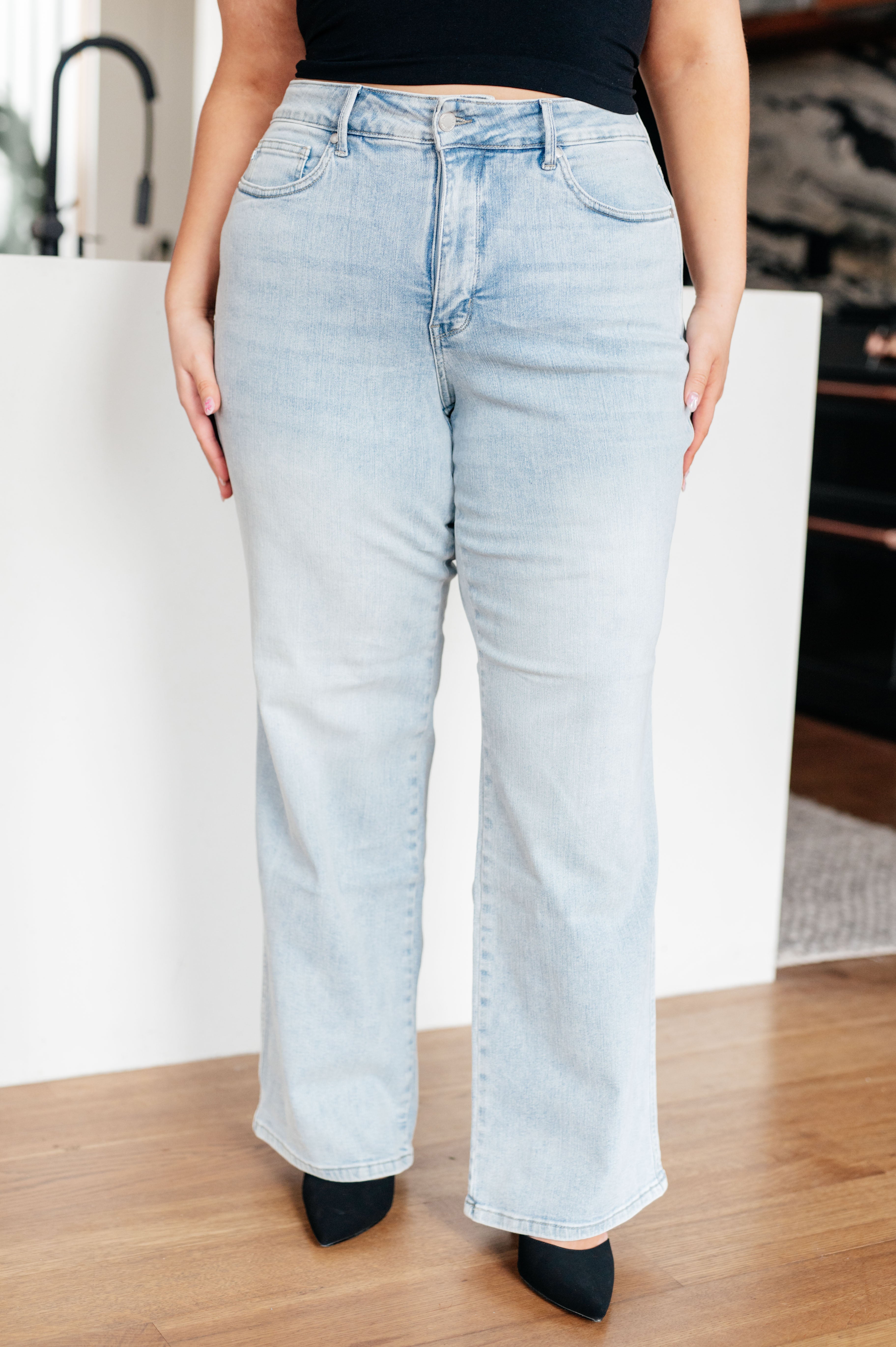 A person with long brown hair is standing in a modern kitchen, wearing a black sleeveless top and light blue Brooke High Rise Control Top Vintage Wash Straight Jeans by Judy Blue with one hand in their pocket. The kitchen features dark cabinets, a white island, and wooden floors.