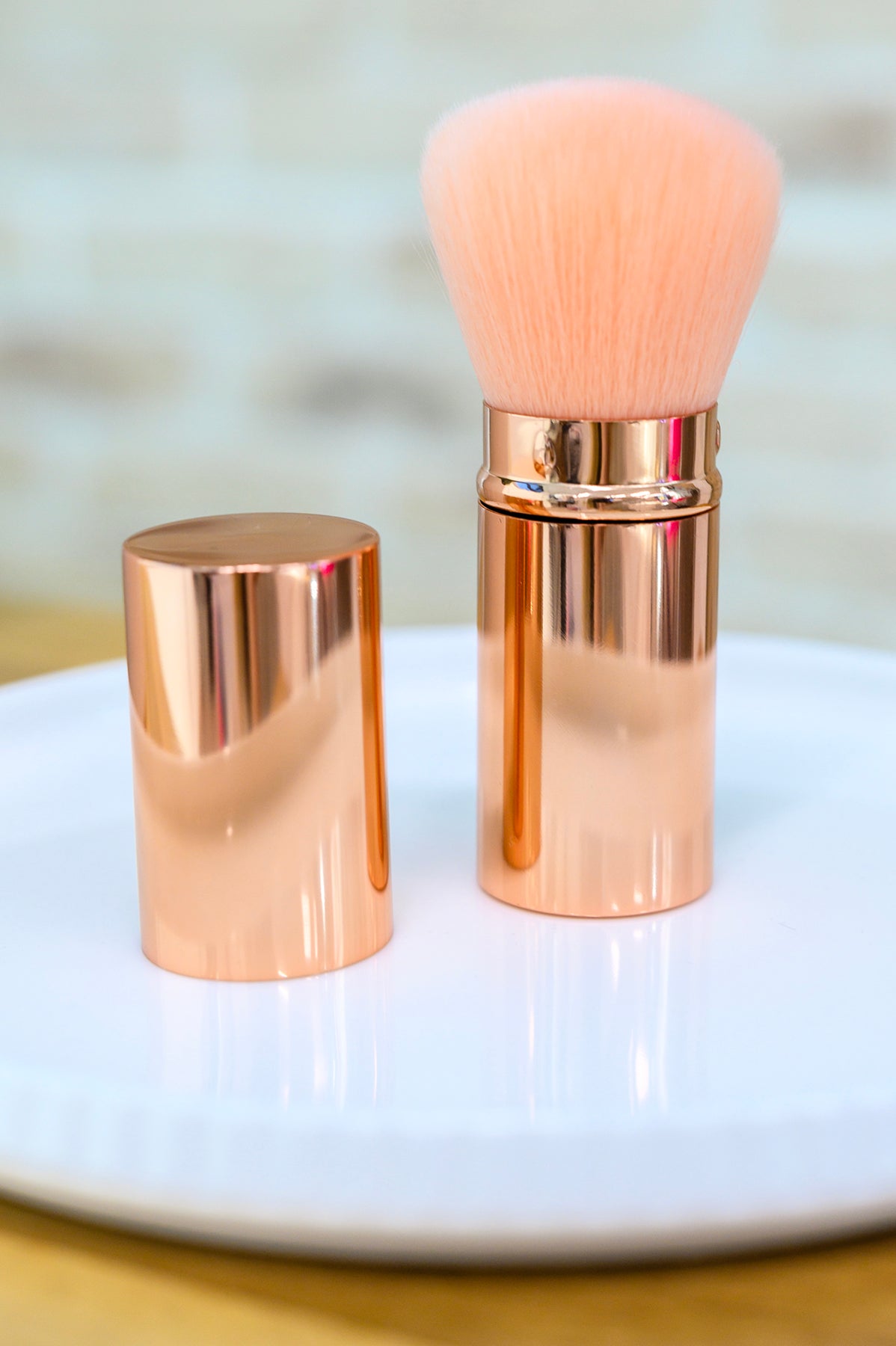 A rose gold Telescopic Powder Brush with a matching cap from The Dapper Squirrel is placed upright on a white plate. The brush, featuring soft, pink synthetic bristles, has the cap positioned next to it. The background is softly blurred, highlighting the brush as the focal point.