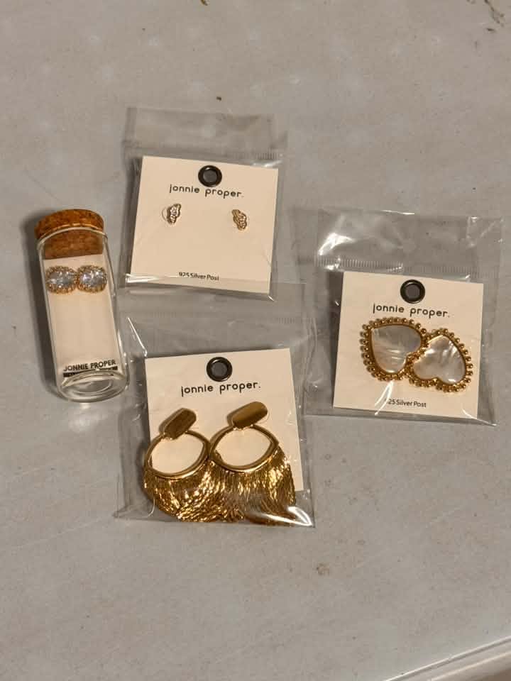 Four pairs of gold and silver jewelry from Jonnie Proper, including earrings and a necklace, displayed on their original white brand cards next to a small glass vial on a gray surface.