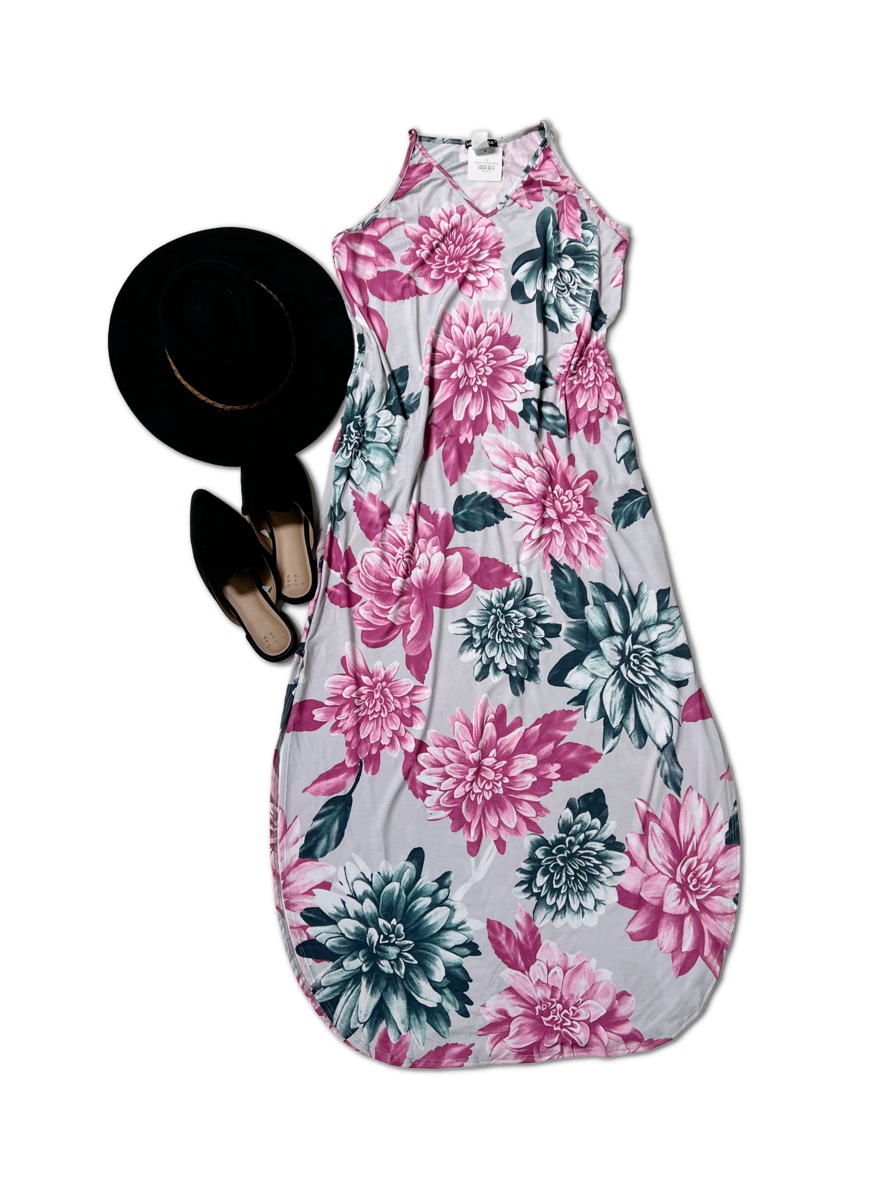 A Blissful Floral Maxi Dress with pink and green flowers on white Heimish fabric is paired with a wide-brimmed black hat and stylish, practical black slip-on shoes with pointed toes, set against a white backdrop.