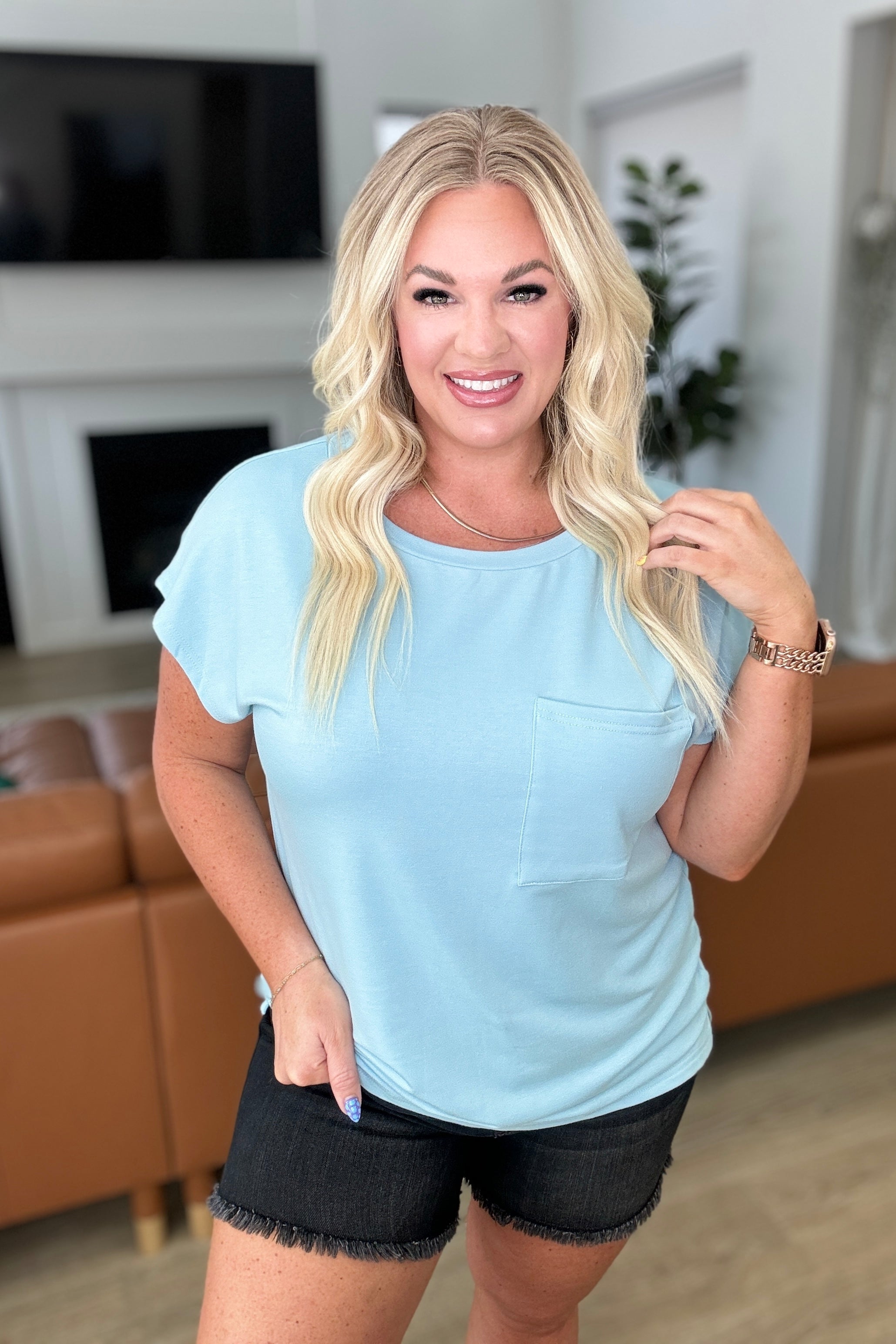 A woman with long blonde hair is smiling while standing indoors. She is wearing a light blue Not So Basic Pocket Tee in Blue by One Eleven North and dark denim shorts. She has a watch on her left wrist, and there is a brown sofa and green plant in the background.