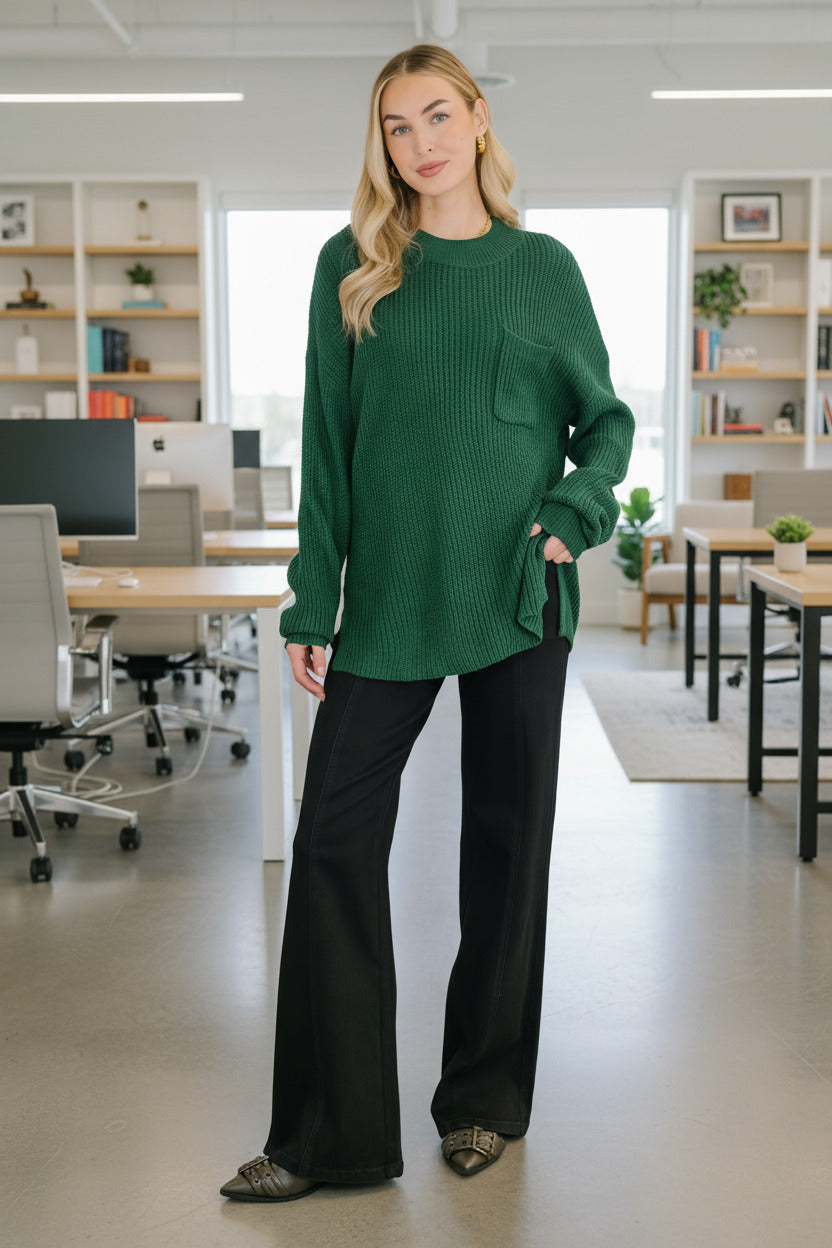Woman wearing a green sweater and black pants standing indoors with a plant and decorative wall in the background.