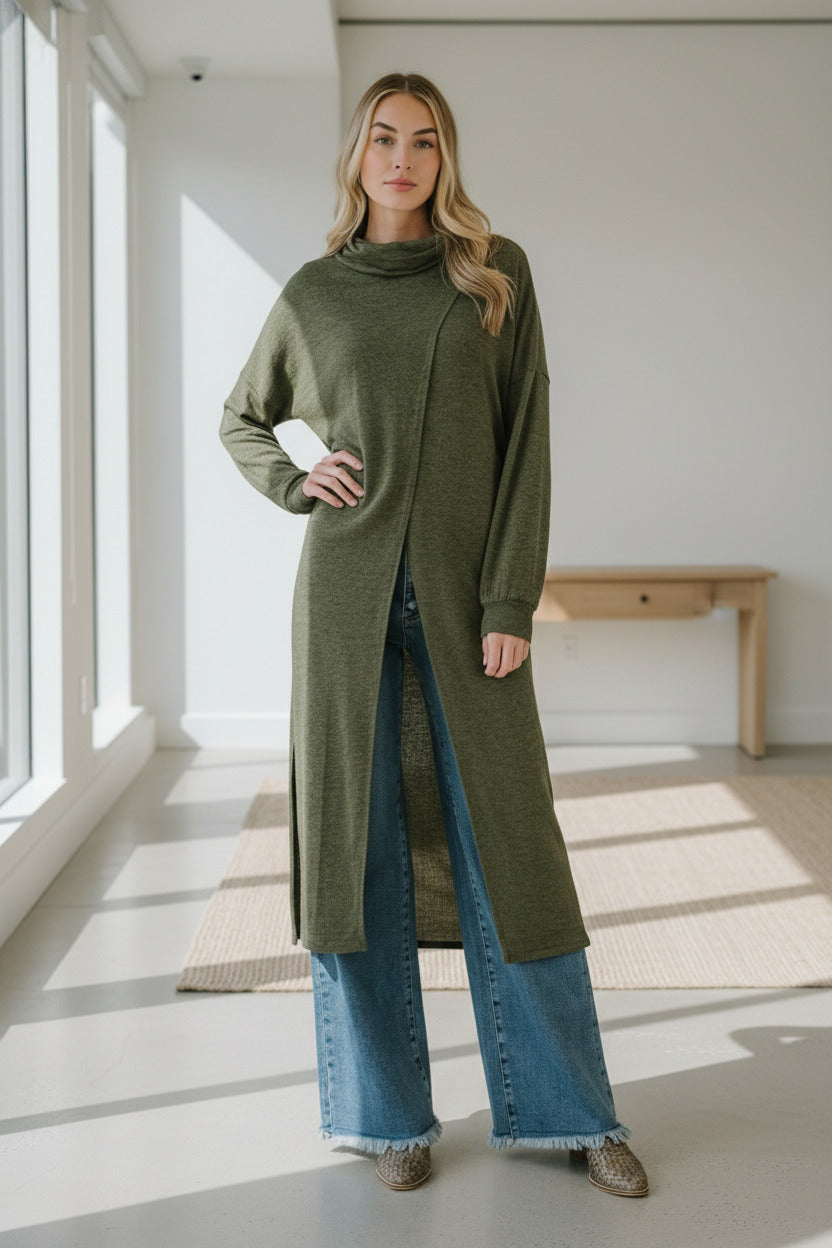 Woman wearing a long olive green sweater with blue jeans indoors.