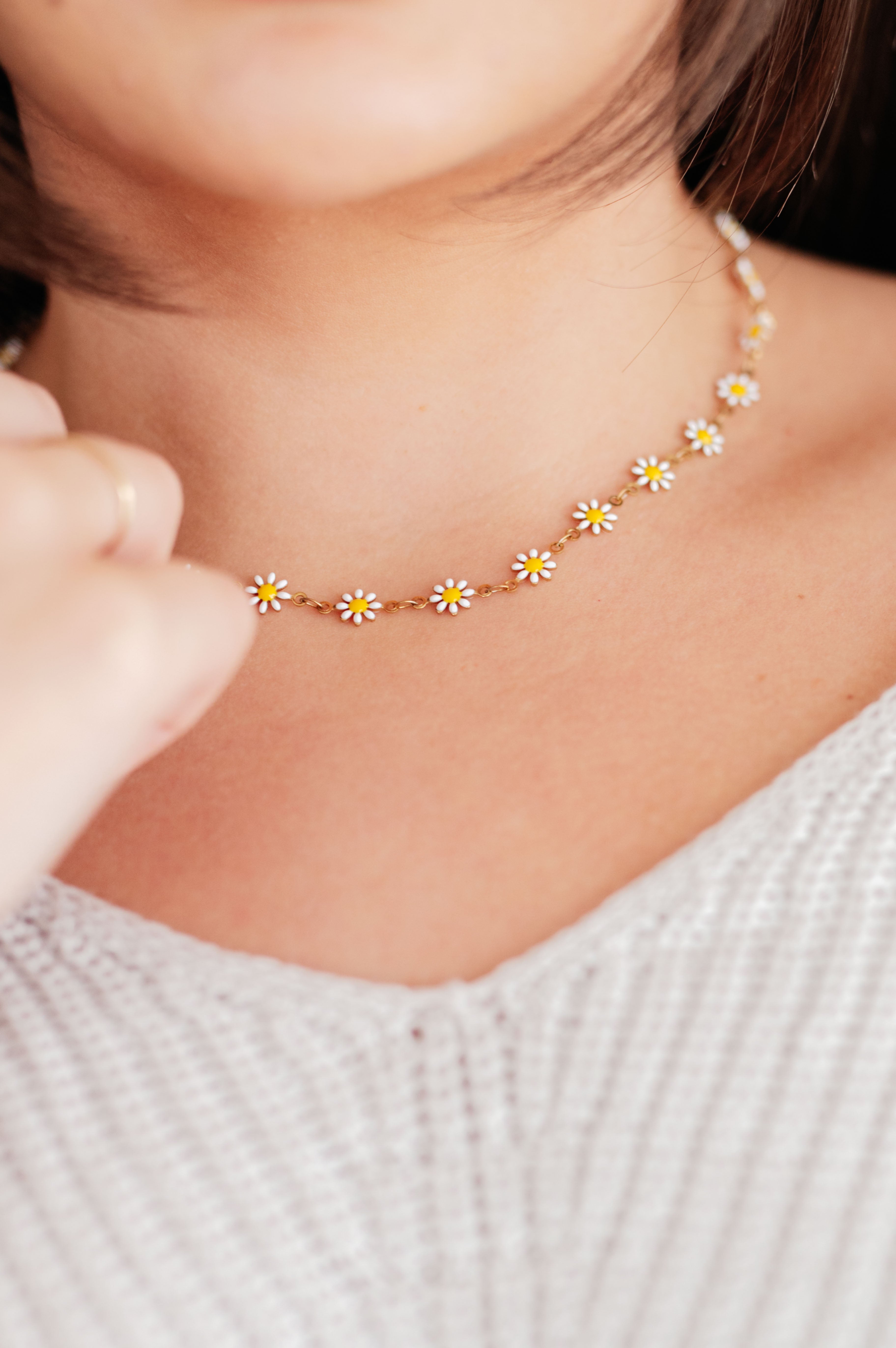 Close-up of a person wearing a delicate Wildflower Necklace in White by The Dapper Squirrel, featuring small white and yellow flower charms. Crafted with 18k gold plated details, this hypoallergenic piece beautifully complements their short, dark hair and light knit sweater. Focus remains on the necklace and the person's neckline.