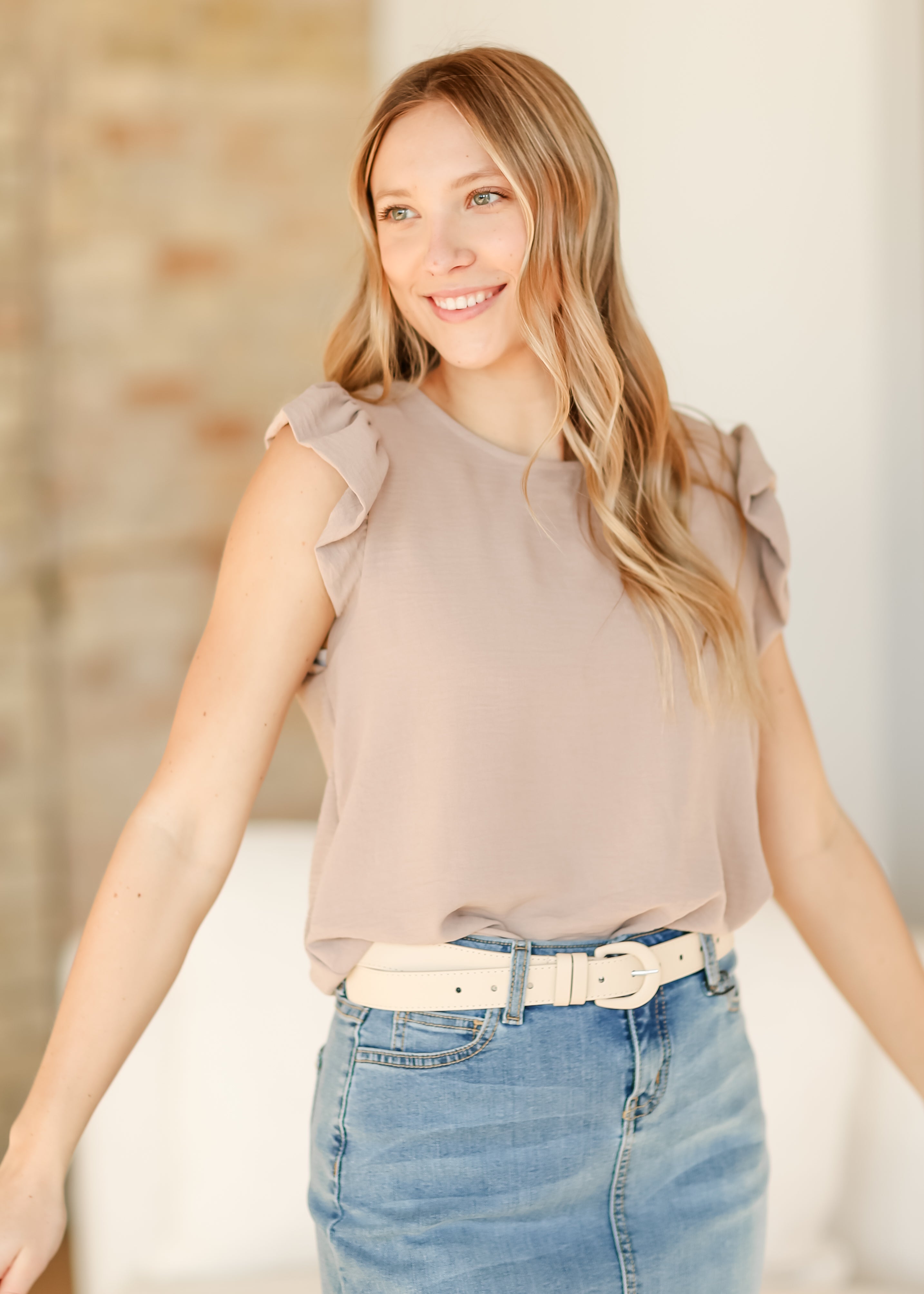 Woman wearing a beige blouse and blue jeans with a white belt, standing against a neutral background.