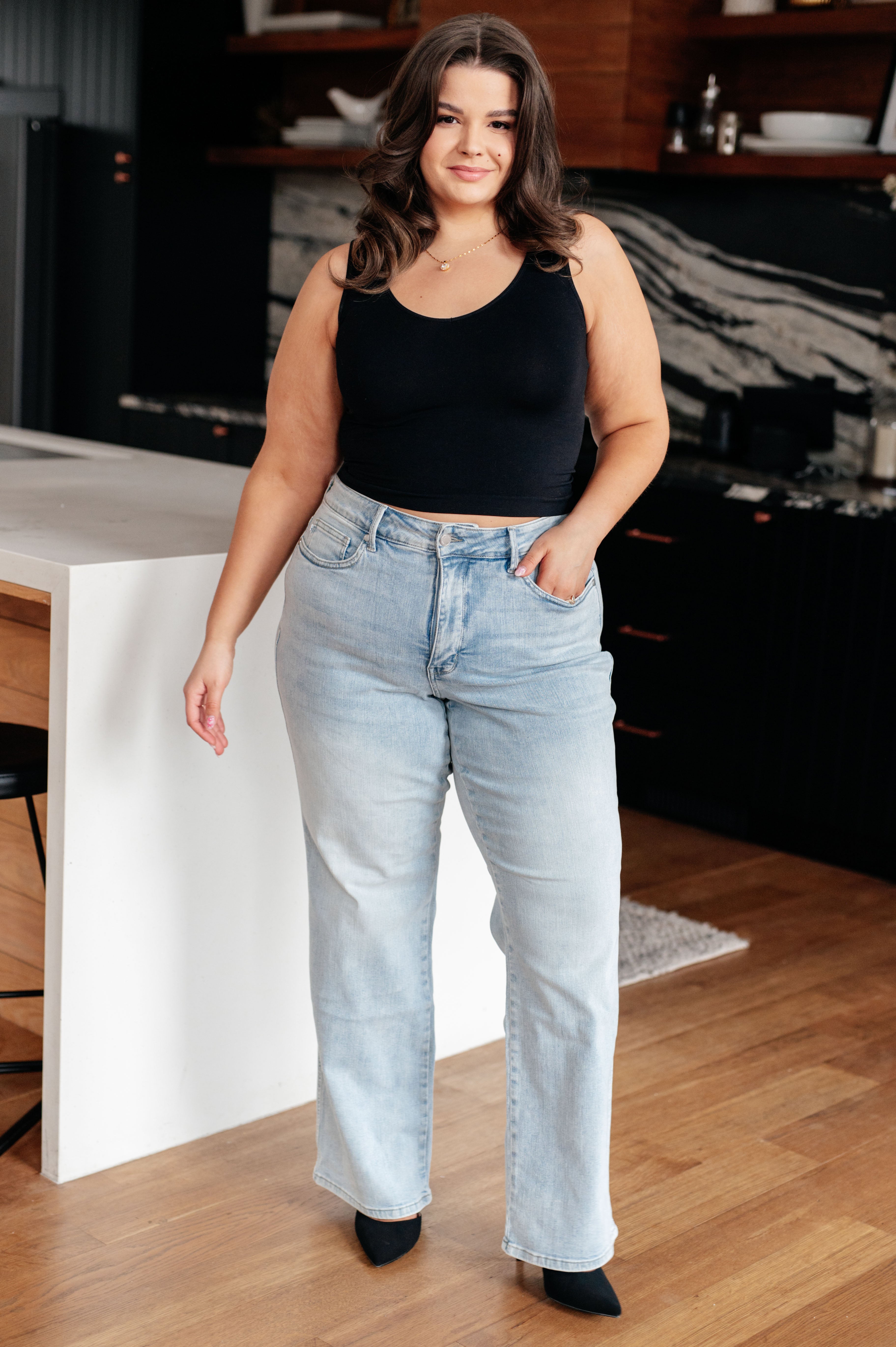 A person with long brown hair is standing in a modern kitchen, wearing a black sleeveless top and light blue Brooke High Rise Control Top Vintage Wash Straight Jeans by Judy Blue with one hand in their pocket. The kitchen features dark cabinets, a white island, and wooden floors.